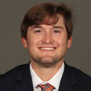 Smiling man wearing an orange tie and black coat in front of a grey background